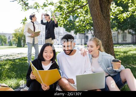 Studente asiatico che tiene notebook vicino agli amici con computer portatile e caffè nel parco Foto Stock