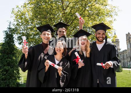 Sorridenti bachelor multiculturali che tengono diplomi e guardano la macchina fotografica nel parco Foto Stock
