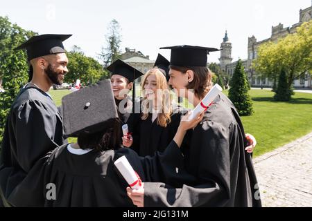 Sorridenti bachelor interrazziali in camici con diplomi e abbraccio in parco Foto Stock
