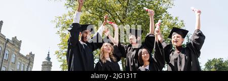 Vista ad angolo basso di eccitati bachelor multietnici in CAPS che tengono diplomi in parco, banner Foto Stock