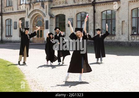 Bachelor eccitato con diploma che mostra sì gesto vicino amici interrazziali e università all'aperto Foto Stock