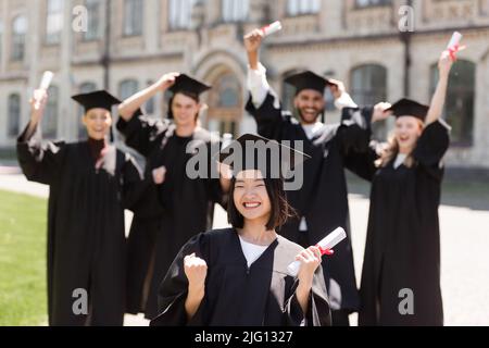 Eccitati bachelor asiatici in possesso di diploma vicino blurred amici nel parco Foto Stock