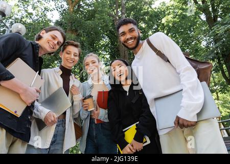 Vista ad angolo basso di studenti multiculturali allegri con notebook e computer portatili che guardano la macchina fotografica nel parco Foto Stock