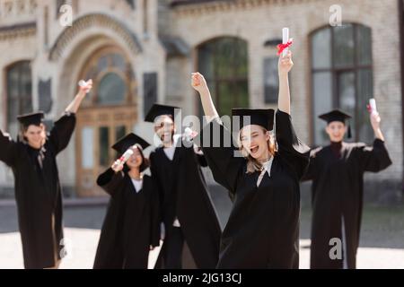 Allievo eccitato in abito di bachelor che tiene il diploma vicino gli amici multietnici all'aperto Foto Stock