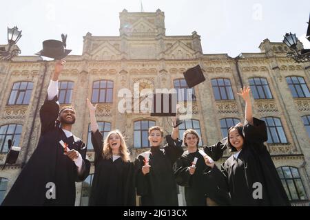 Vista ad angolo basso di felici bachelor multietnici che lanciano tappi vicino all'università all'aperto Foto Stock