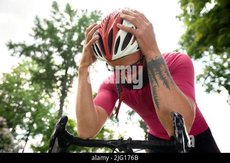 Ciclista maschile stanco in abbigliamento sportivo, occhiali e casco protettivo camminare con la sua bicicletta nella foresta per fare una pausa dopo la guida. Blu cielo e foresta Foto Stock