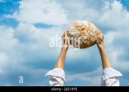 Le mani femminili tengono la pagnotta di pane fatta in casa sopra la sua testa sopra un cielo blu di estate. Concetto di sicurezza alimentare mondiale. Foto Stock