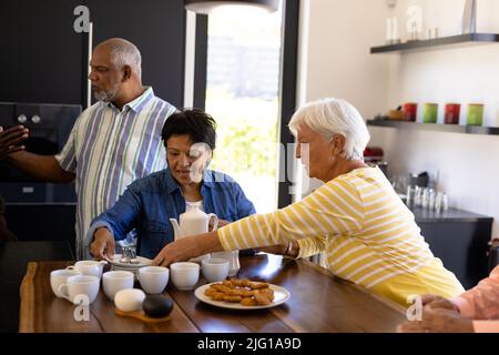 Donne anziane biraciali che servono caffè e biscotti agli amici su tavola di legno in casa di cura Foto Stock