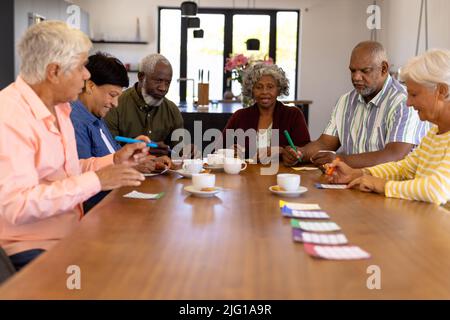 Amici senior multirazziali che giocano a bingo mentre hanno caffè e biscotti al tavolo da pranzo Foto Stock