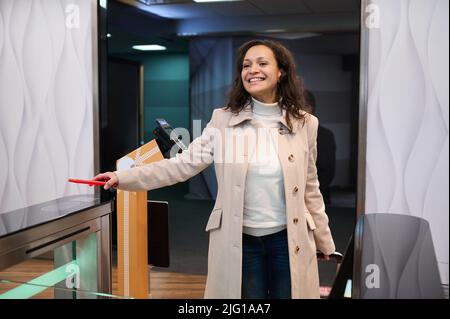 Sorridente donna multietnica bella, in abbigliamento casual in un viaggio di lavoro, scansiona le informazioni sul suo smartphone, lo posiziona sul lettore di codice QR al Foto Stock