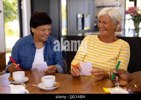 Allegro biraciale senior femminile amici con caffè e biscotti sul tavolo da pranzo giocare a bingo Foto Stock