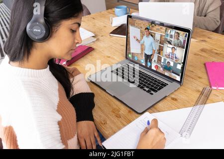 Ragazza seria di adolescenza biraciale che scrive nel libro mentre imparano in linea sopra la videochiamata sul laptop Foto Stock