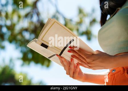 Bella donna brunetta con t-shirt turchese in piedi sul parco della città, all'aperto lettura libro istruzione studiare conoscenza di apprendimento. Foto Stock