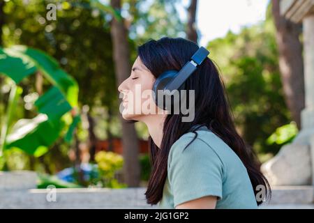 Vista laterale della donna millenaria carina con tee turchesi e cortine arancioni sul parco cittadino, all'aperto ascoltando musica con cuffie e occhi chiusi. Divergetevi Foto Stock