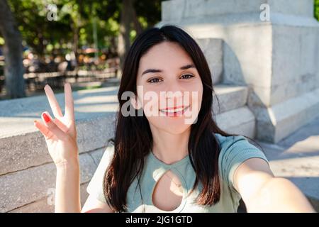 Giovane ragazza brunette sorridente felice indossare t-shirt turchese in piedi sul parco della città, all'aperto sorridendo con il viso felice winking allo smart phone facendo v Foto Stock