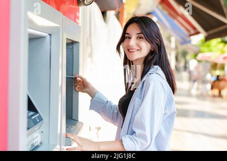 Donna bruna carina che indossa una maglietta in piedi sul parco cittadino, all'aperto davanti al bancomat, sorridente e con carta di credito o debito. In cerca Foto Stock