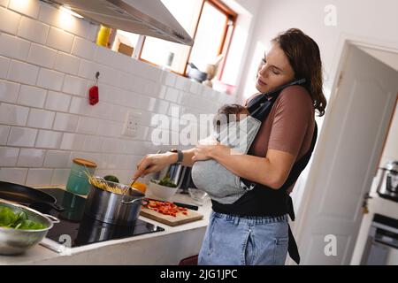 Metà adulto caucasico madre rispondere telefono e portare neonato bambino mentre cucinando cibo in cucina Foto Stock