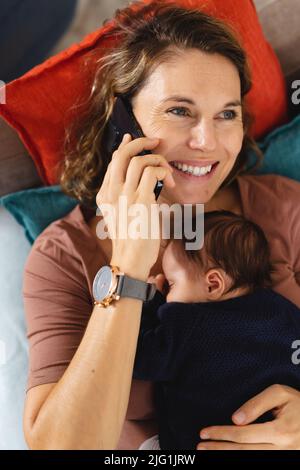 Sorridente madre caucasica adulto medio che risponde smartphone con il neonato che dorme sul divano Foto Stock