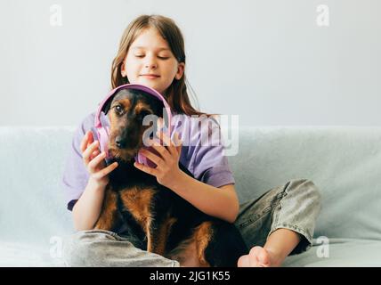 Ragazza felice adolescente in t-shirt lilla siede sul divano, ascolta la musica ha divertimento giocando con il cane Foto Stock