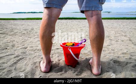 Gli uomini adulti sono in piedi all'aperto sulla spiaggia con secchio di plastica, auto giocattolo in legno con pala giocattolo all'interno Foto Stock