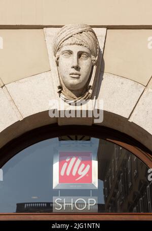 TRIESTE, Italia - 25 marzo 2022: Scultura neoclassica di testa femminile sulla facciata di un edificio storico sopra una caffetteria con il logo del caffè Illy Foto Stock