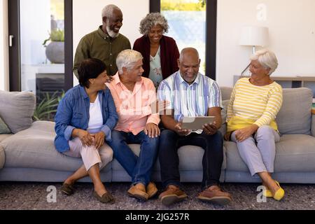 Allegri amici senior multirazziali che guardano l'uomo anziano utilizzando un tablet digitale mentre si siede sul divano Foto Stock