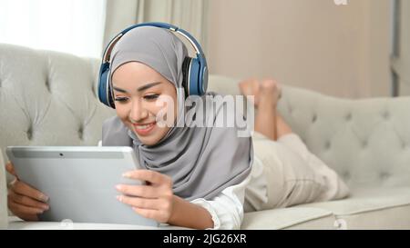 Donna musulmana asiatica rilassata e felice con hijab che si stende sul divano, ascoltando musica con le cuffie e utilizzando il tablet digitale. Foto Stock
