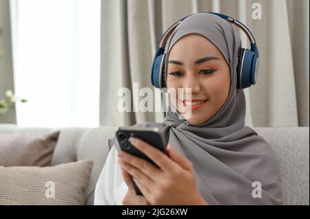 Donna musulmana asiatica dal taglio millenario con hijab in salotto, ascoltando musica con cuffie wireless e utilizzando il suo smartphone. Foto Stock
