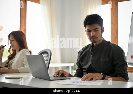 Bell'uomo d'affari asiatico giovane-adulto e intelligente si è concentrato a lavorare su un rapporto di dati finanziari e utilizzando il computer portatile in ufficio. Foto Stock