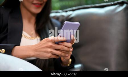 Splendida donna asiatica o donna d'affari si rilassa sul divano e utilizza uno smartphone moderno per inviare messaggi a qualcuno. Immagine ritagliata Foto Stock