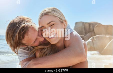 Coppia felice e amorevole che si gode una giornata in spiaggia in estate. Il marito allegro abbraccia la moglie gioiosa mentre si trova in posizione sul mare in vacanza Foto Stock