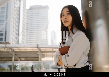 Ritratto di bella giovane donna asiatica in piedi appoggiata a pilastro edificio tenendo un notebook e penna mentre si pensa a qualcosa e guardando via Foto Stock