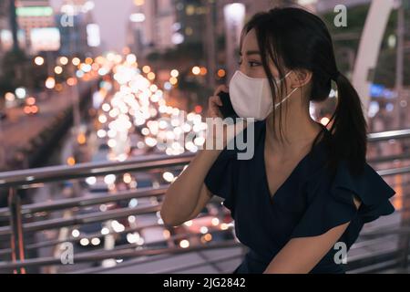 Donna asiatica giovane e intelligente che indossa la maschera di sicurezza COVID-19 seduta e in attesa di qualcuno e parlare al telefono in città strada durante la sera, mentre si sente sconvolto Foto Stock