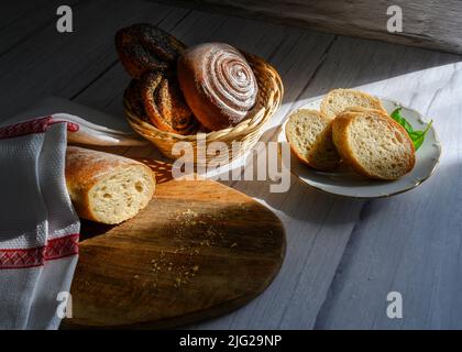 Pane sotto un asciugamano su un asse di legno e in un piatto, ciambelle in un cestino di vimini Foto Stock