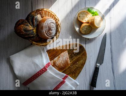 Pane sotto un asciugamano su un asse di legno e in un piatto, ciambelle in un cestino di vimini e un coltello Foto Stock