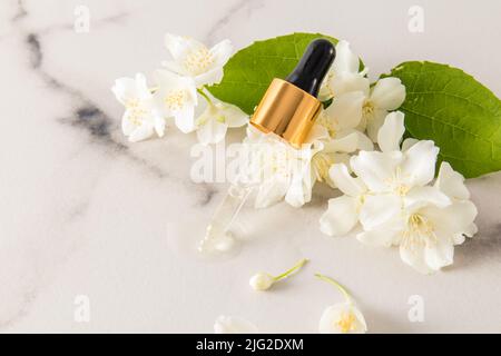 Il contagocce di una bottiglia cosmetica con olio naturale di gelsomino si trova sui fiori. Spazio per il testo. Sfondo bianco marmo Foto Stock