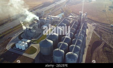 Volo aereo con vista sui droni sopra il complesso di edifici di ascensore di grano. Silos rotondi in metallo e barili in metallo per la conservazione dei cereali. Raccolta di stoccaggio in essiccazione. Serbatoi dell'elevatore e complesso di essiccazione della granella. Granaio grande Foto Stock