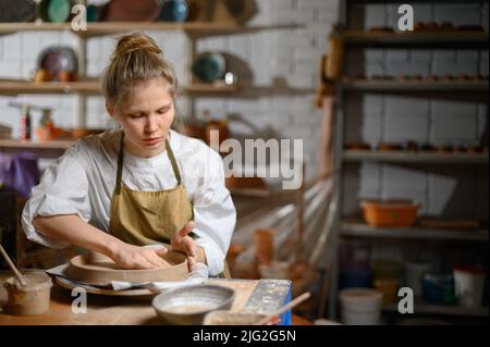 Un ceramista fa un piatto. Donna in grembiule lavora in un laboratorio di ceramica. Foto Stock