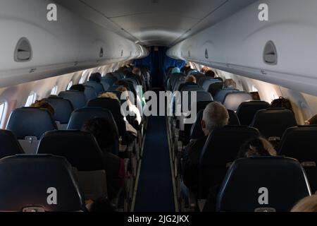 Vista dall'interno dell'aereo passeggeri cabina e corridoio dal retro. Le persone sedute su sedili aerei e i compartimenti per la testa chiusi sopra di loro Foto Stock