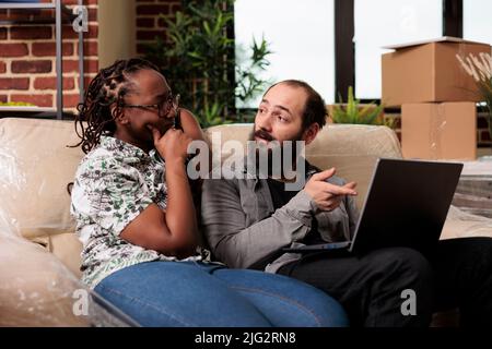 Diversi partner di vita facendo shopping online su laptop per acquistare mobili e decorare nuova casa. Muovendosi nella nuova proprietà dell'appartamento sul prestito, trovando l'ispirazione del decor sul Web site del Internet. Foto Stock