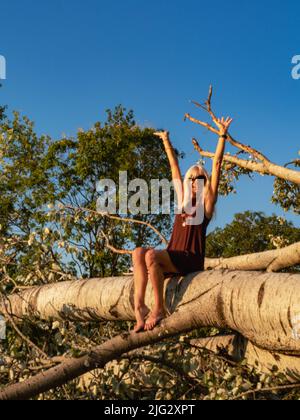 donna bionda caucasica adulta alta in un parco vicino ad un grande albero caduto. Ritratto all'aperto di donna matura sicura e positiva. Sorridente bionda femmina Foto Stock