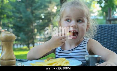 La bambina mangia patatine fritte. Primo piano di ragazza bionda prende patatine con le mani e li prova seduti in strada caffè sul parco. Foto Stock