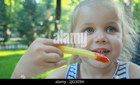 La bambina mangia patatine fritte. Primo piano di ragazza bionda prende patatine con le mani e li prova seduti in strada caffè sul parco. Foto Stock