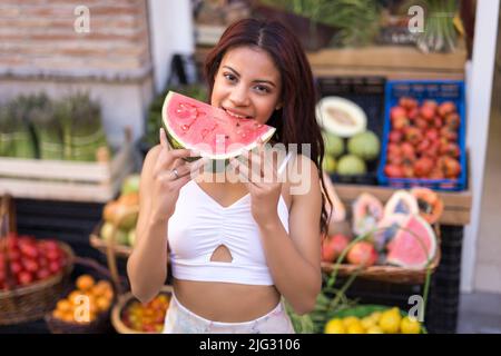 Positiva giovane donna in abiti casual sorridente e guardando la macchina fotografica mentre si trova vicino agli scaffali con verdure assortite e frutta Foto Stock