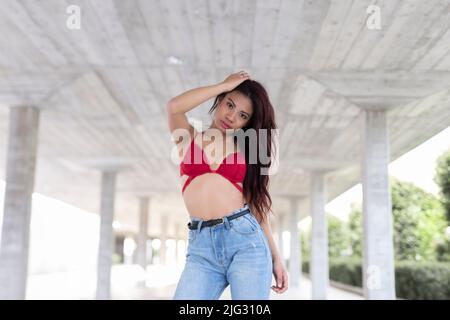 Basso angolo di giovane etnia femminile in abito alla moda che tocca i capelli e guarda la macchina fotografica mentre si trova sotto il ponte in città Foto Stock