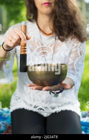 Dettaglio delle mani di una donna che tiene e suona una ciotola di canto tibetano durante una sessione di meditazione e terapia musicale in un parco. Foto Stock