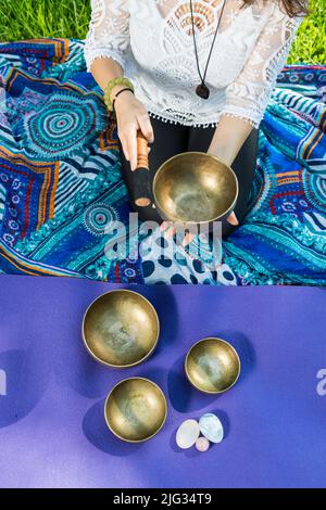 Vista dall'alto di una donna che tiene e suona una ciotola di canto tibetano durante una sessione di meditazione e di terapia musicale su un tappetino yoga. Foto Stock