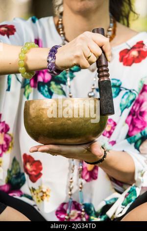 Dettaglio delle mani di una donna che tiene e suona una ciotola di canto tibetano durante una sessione di meditazione e terapia musicale in un parco. Foto Stock