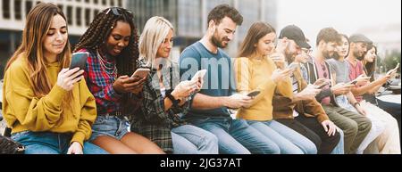 Gen z persone diverse utilizzando cellulari seduti su una panca all'aperto, panorama. Concetto di stile di vita con uomini e donne multirazziali utilizzando smartphone. Gadge Foto Stock