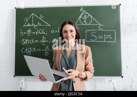 Insegnante sorridente che tiene gesso e laptop vicino a formule matematiche sulla lavagna Foto Stock